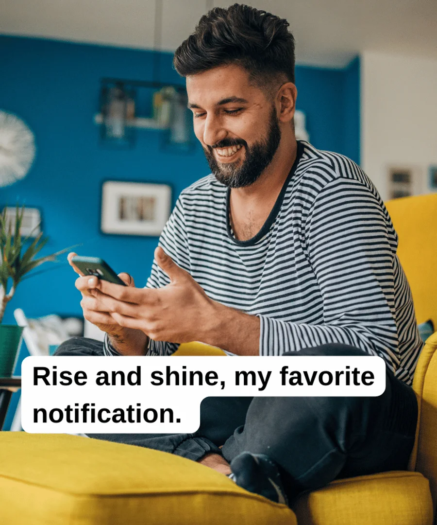 A man sitting on a yellow sofa smiling at a message from his crush. The text reads: “Rise and shine, my favorite notification.”
